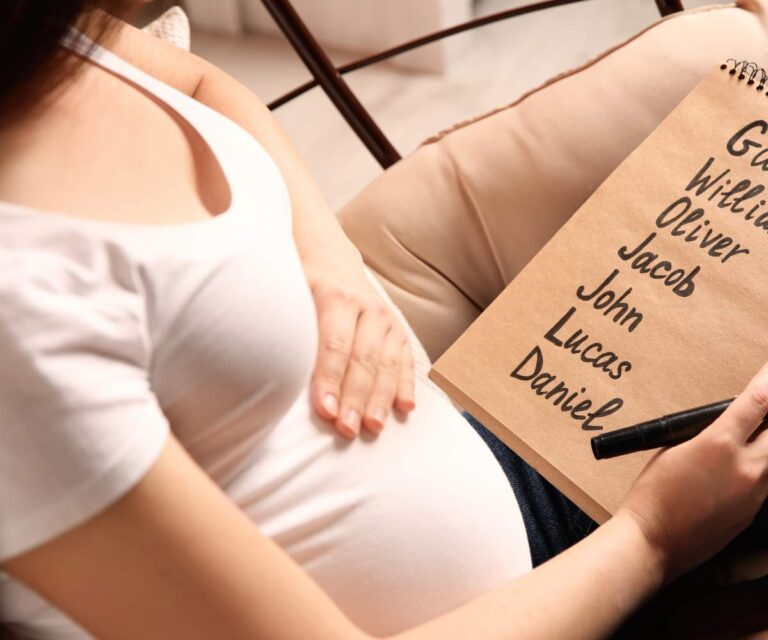 Na zdjęciu widoczna jest kobieta w ciąży, siedząca na krześle i trzymająca na kolanach notes. Na notesie, wypisane czarnym markerem, znajdują się popularne imiona męskie. Prawdopodobnie kobieta zastanawia się nad wyborem imienia dla chłopca